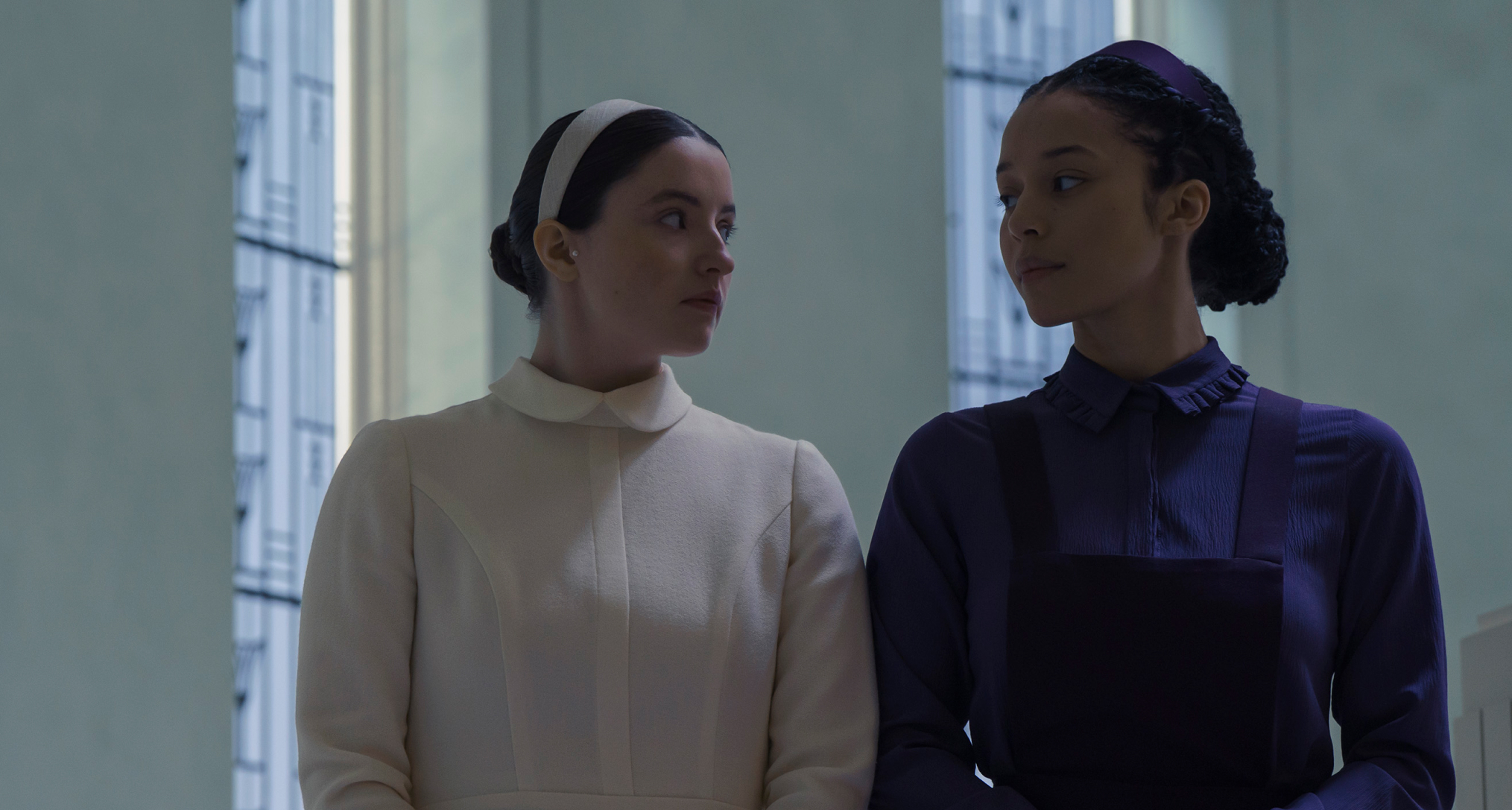 lucy halliday as daisy in white and chase infiniti as agnes in purple walking through their school hallways in the testaments