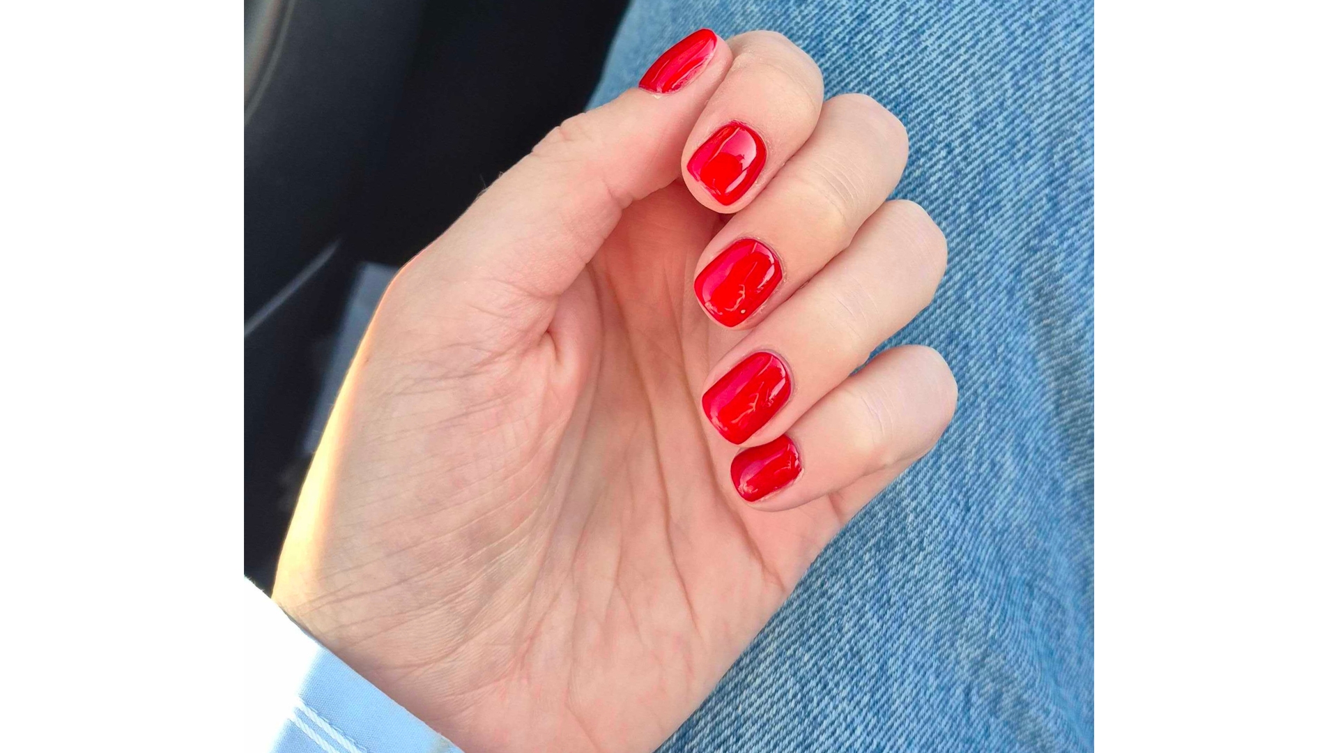 A close-up of Digital Beauty Writer, Naomi Jamieson&#039;s hand, seen with short and square bright red nails/ in front of a blue-denim background