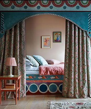 A double four poster bed with colourful bedding and heavy curtains and pelmet. Layers of material, color and pattern to create a cozy nook bed