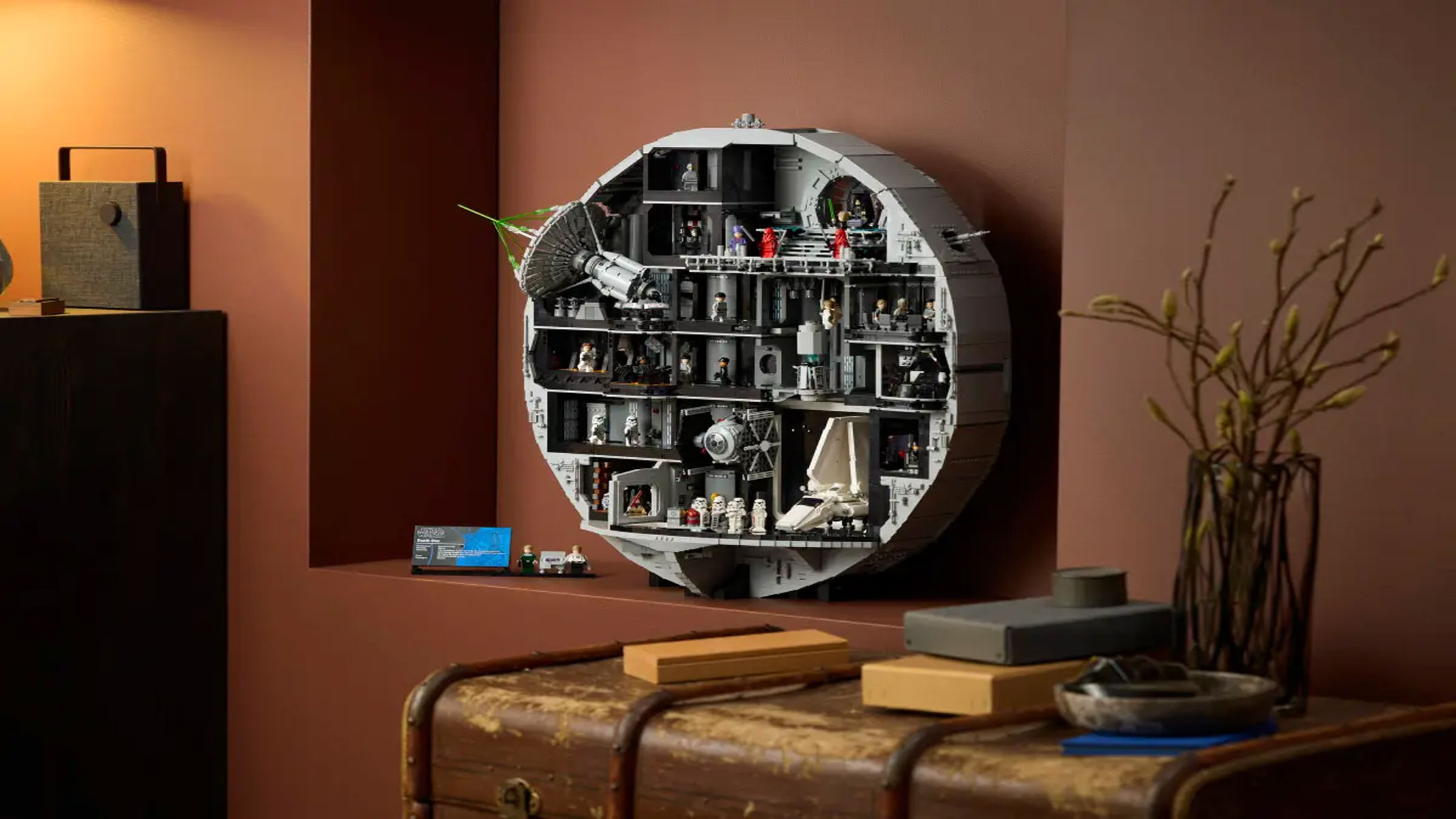 The circular interior of a space station is on show with several floors and rooms filled with minifigures, all in front of a brown wall with a houseplant on the right hand side.