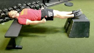 Man performing a Chinese plank with upper back supported on a bench and heels supported on box during gym workout