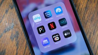 A grid of streaming service apps (Sling, Prime Video, Paramount Plus, Peacock, Netflix, AMC Plus, HBO Max, Pluto TV, Apple TV) open on a phone