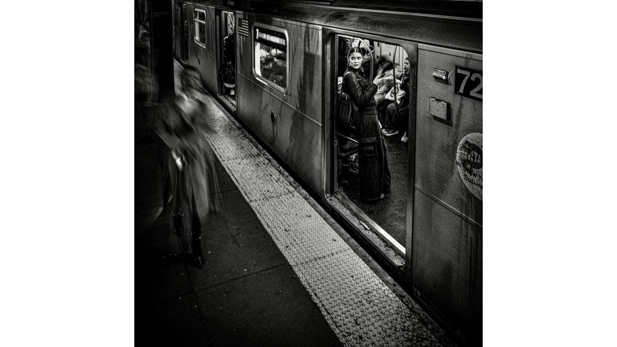 Black and white photo of a subway train with a woman standing in the open door looking out while blurred figures move on the platform