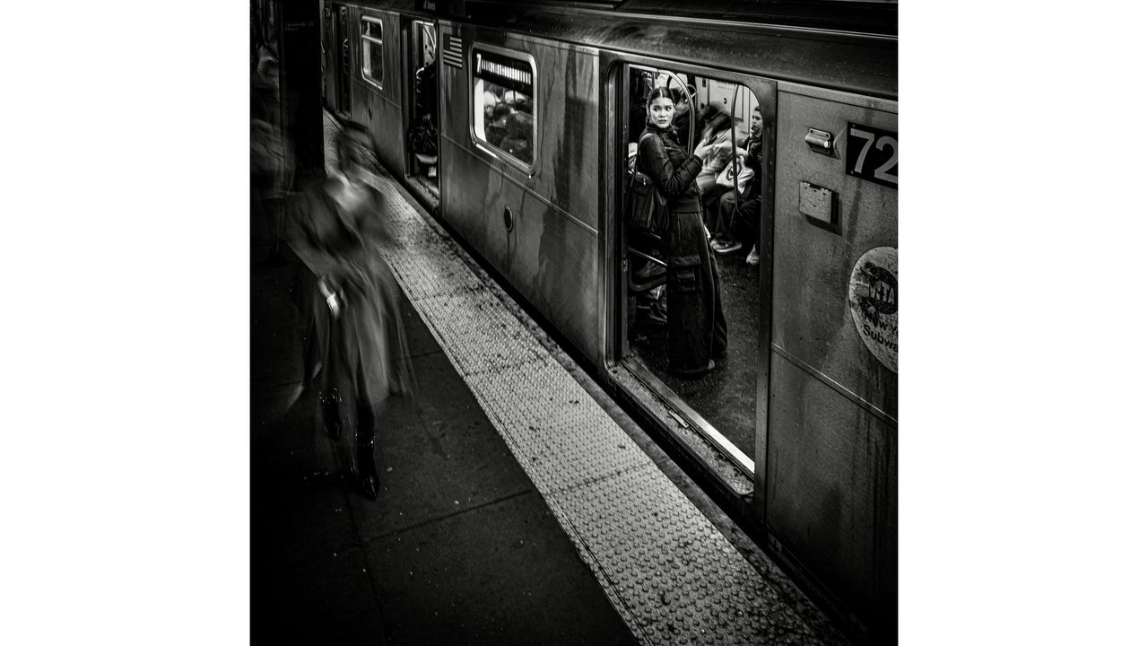 Black and white photo of a subway train with a woman standing in the open door looking out while blurred figures move on the platform