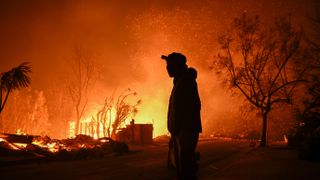 A photo of a person standing in front of a burning house in Pacific Palisades, Los Angeles.