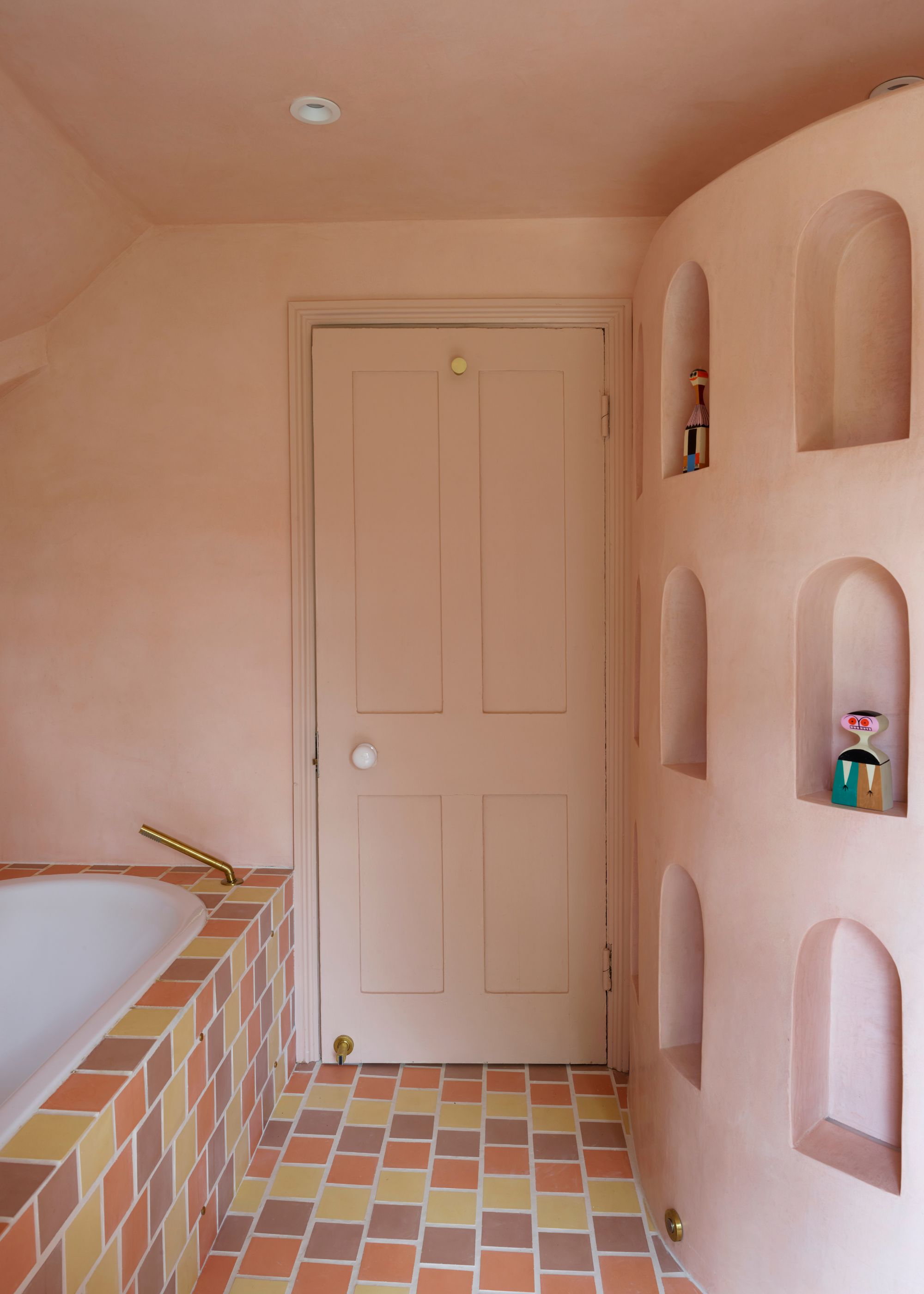 A modern bathroom with plaster pink color-drenched walls and a matching door, pink, yellow, and orange floor tiles continued to the side of the bath, and arched recessed shelving on the wall.