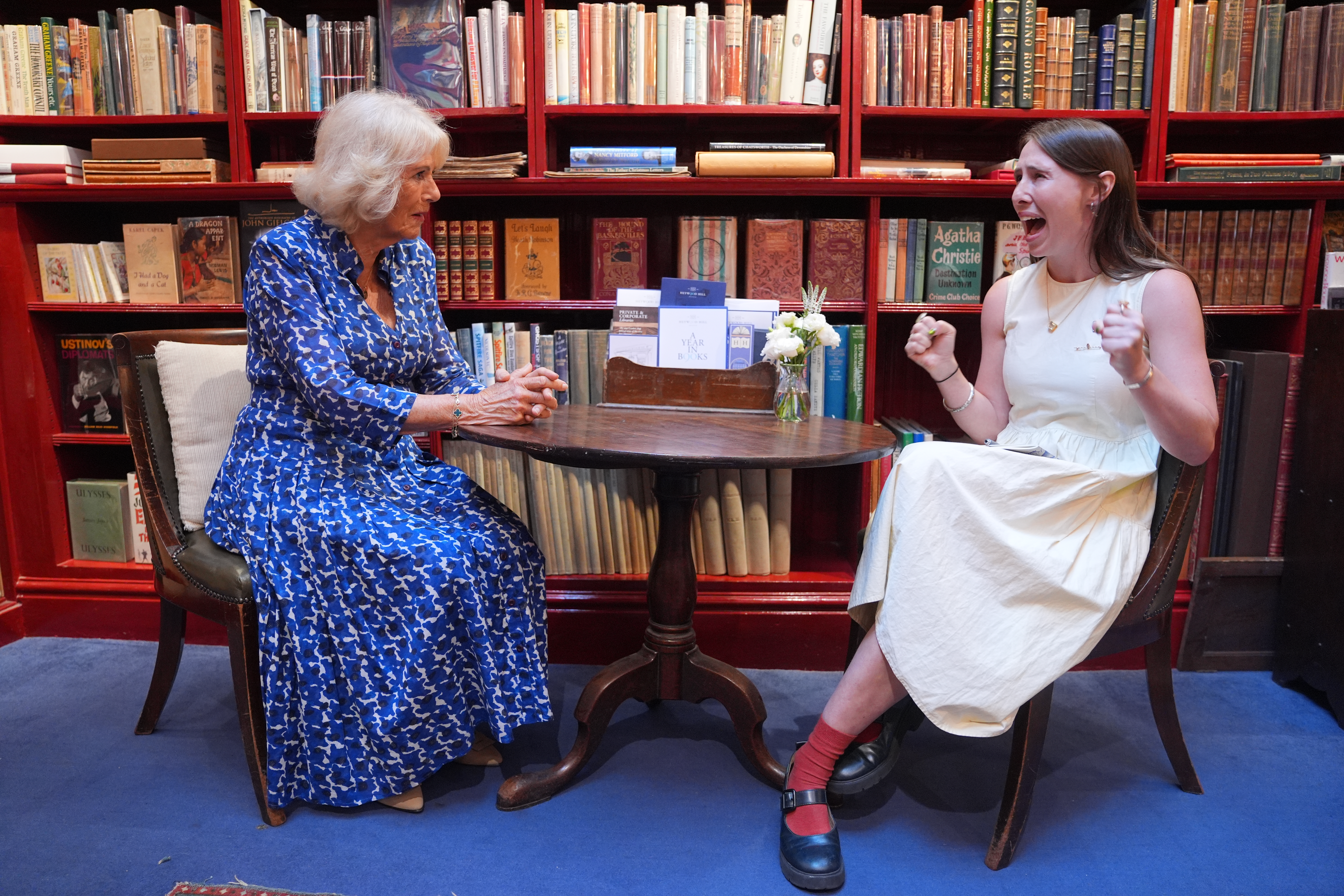 Queen Camilla sitting at a table talking to a woman who is reacting in shock