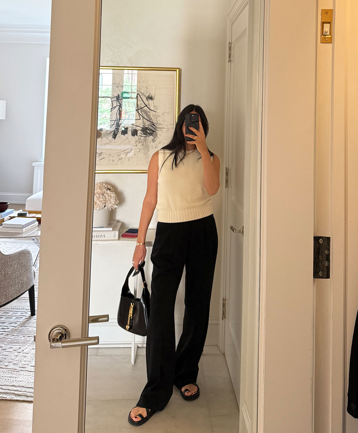 @nicoleakhtarzad taking a mirror outfit selfie and wearing a sleeveless sweater and trousers.