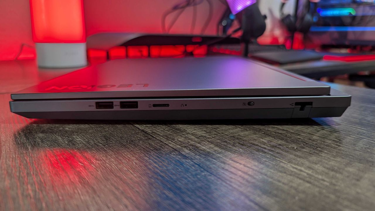 A Lenovo Legion 5i (Gen 9) gaming laptop on a desk with a red backlight, seen from the left to show the two USB Type-A 3.2 Gen 1 ports, the microSD card slot, the electronic front-facing camera privacy shutter, and the RJ45 Ethernet port.