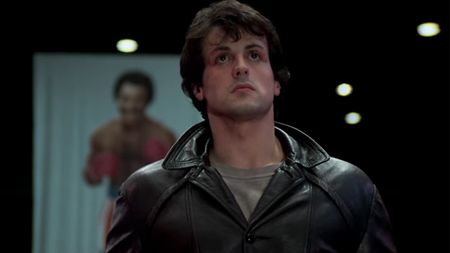 Rocky in a leather jacket with a poster of Apollo behind him