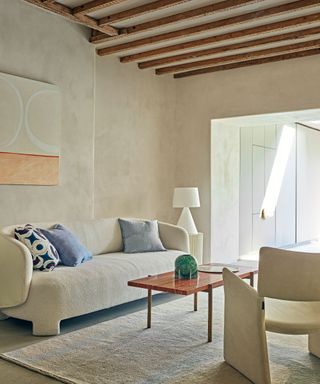 Neutral, relaxed living room scheme with dark wooden ceiling beams, textured cream painted walls, curved boucle sofa with blue scatter cushions, large cream and blue rectangular rug, sculptural cream armchair, dark wooden coffee table, muted geometric artwork, open plan, looking on to other room