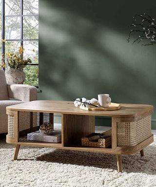 A living room with a dark green wall and a large window, with a light wooden coffee table in the middle with storage space underneath and decor in this and above it