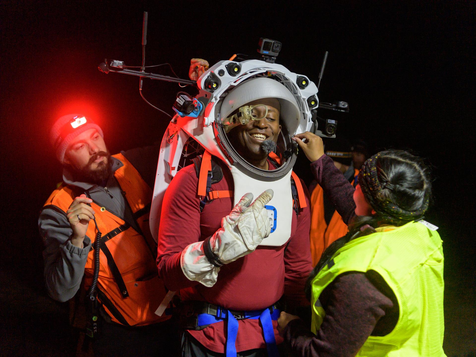 an astronaut at center of the picture wearing a simulated helmet and spacesuit. two engineers on either side adjust his helmet