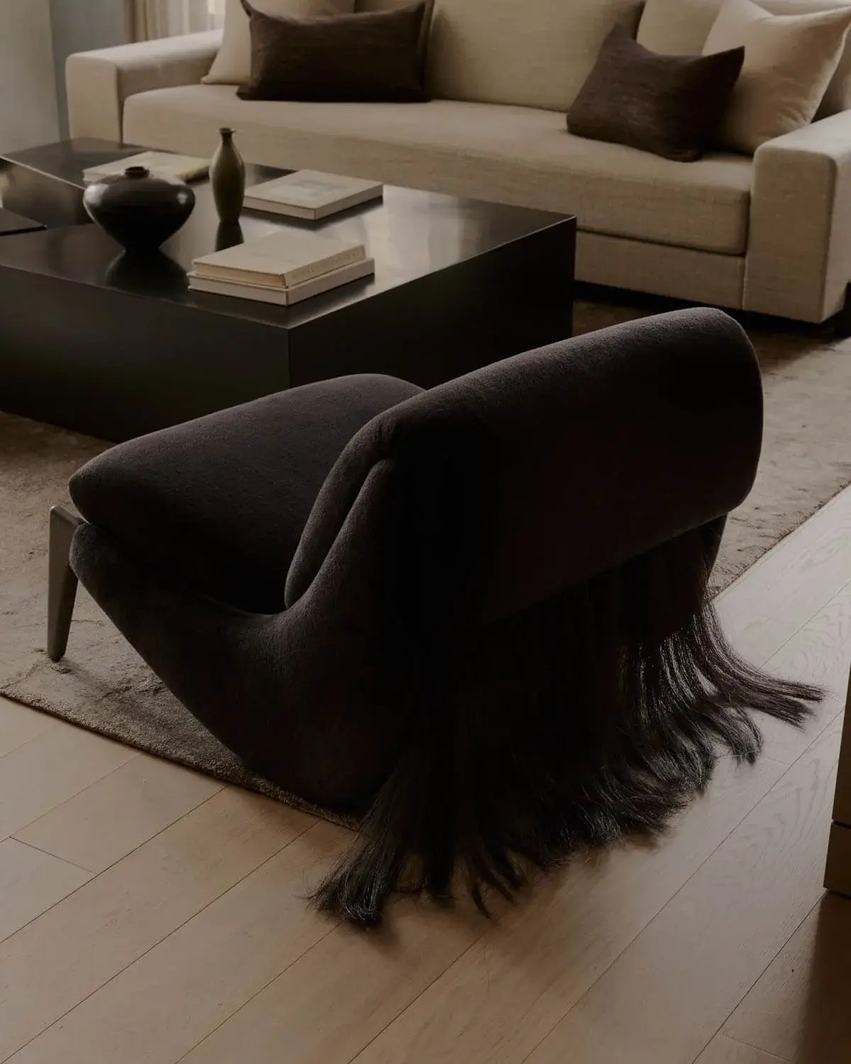 Image of a modern, black accent chair with hair-like long fringe hanging off of the back. The chair is in a modern living room with a large black coffee table, beige rug, and beige sofa in view.