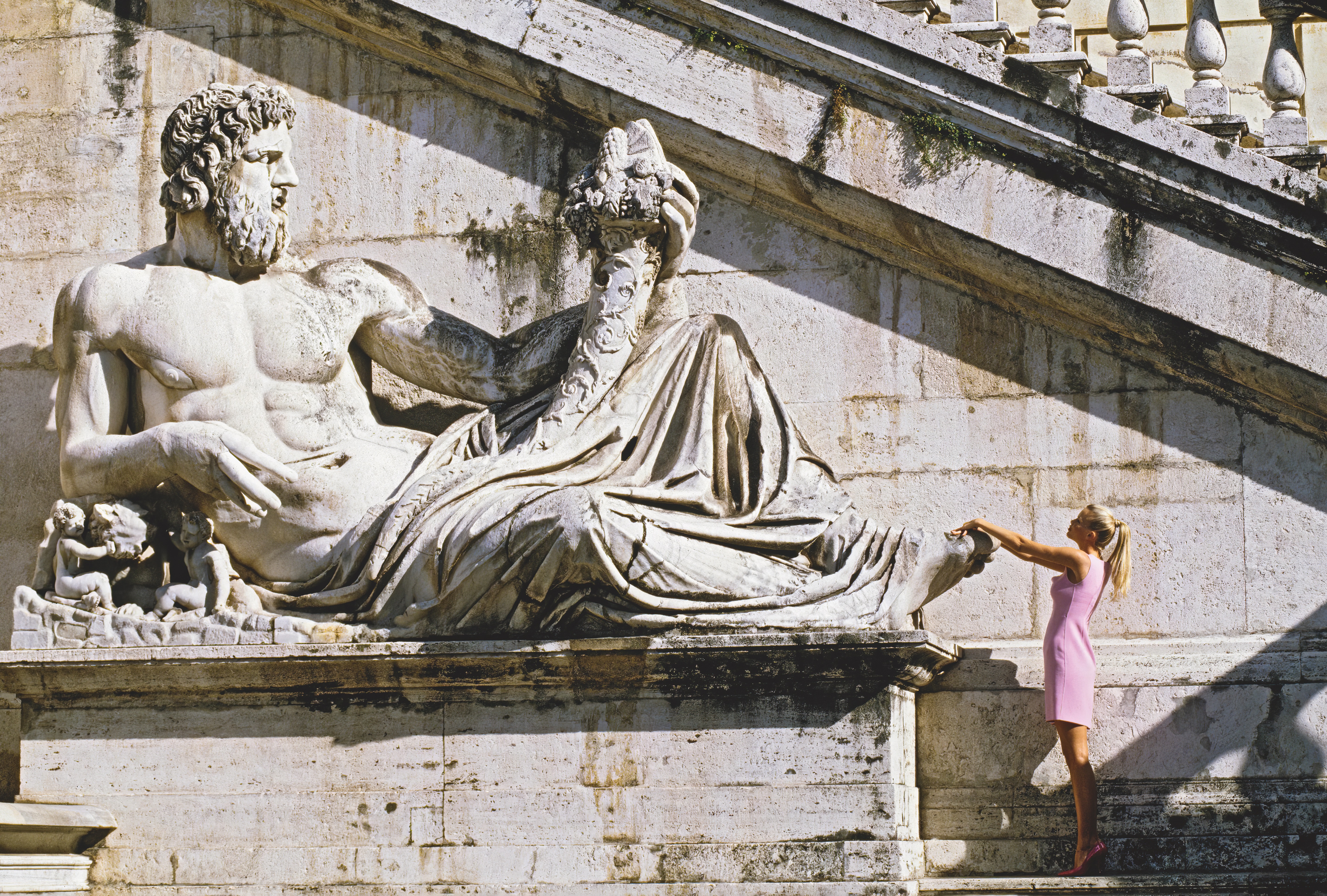 Women in a pink mini dress touching the toe of a giant marble statue