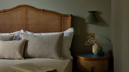 Close-up image of a rattan bed with gray/beige bedding in a sage green bedroom. There is a wooden side table with a green sconce light above it.