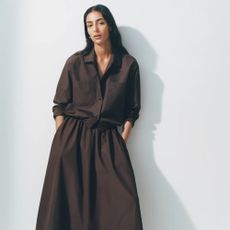 A woman leaning against a white wall while wearing a matching brown shirt and long skirt from ARKET