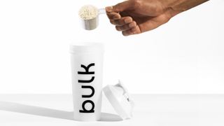 Huel Black Edition vs Bulk 1 Complete Food Shake: person dipping a scoop of Bulk 1 into a shaker placed on a table