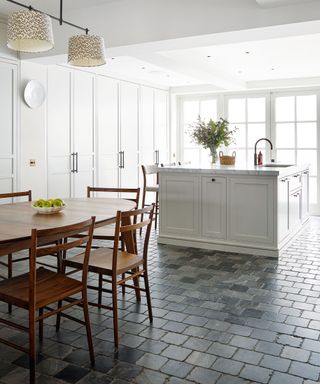 Contemporary kitchen space with white painted walls, cabinets and island, dark gray brick tiled flooring, wooden dining table, rail of lights with shades above dining table