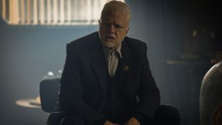 Tim Robbins sits with a puzzled look on his face in a room of low lighting in Silo Season 2.