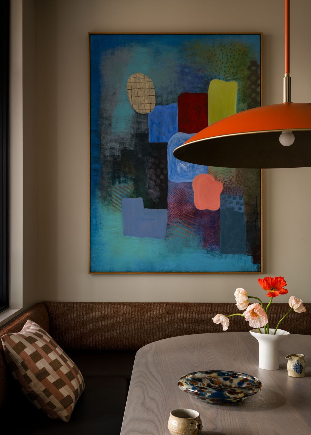 A banquette seating area with colorful artwork above it and a red pendant light above the table with a decorative tray and a small vase with pink and red floral stems