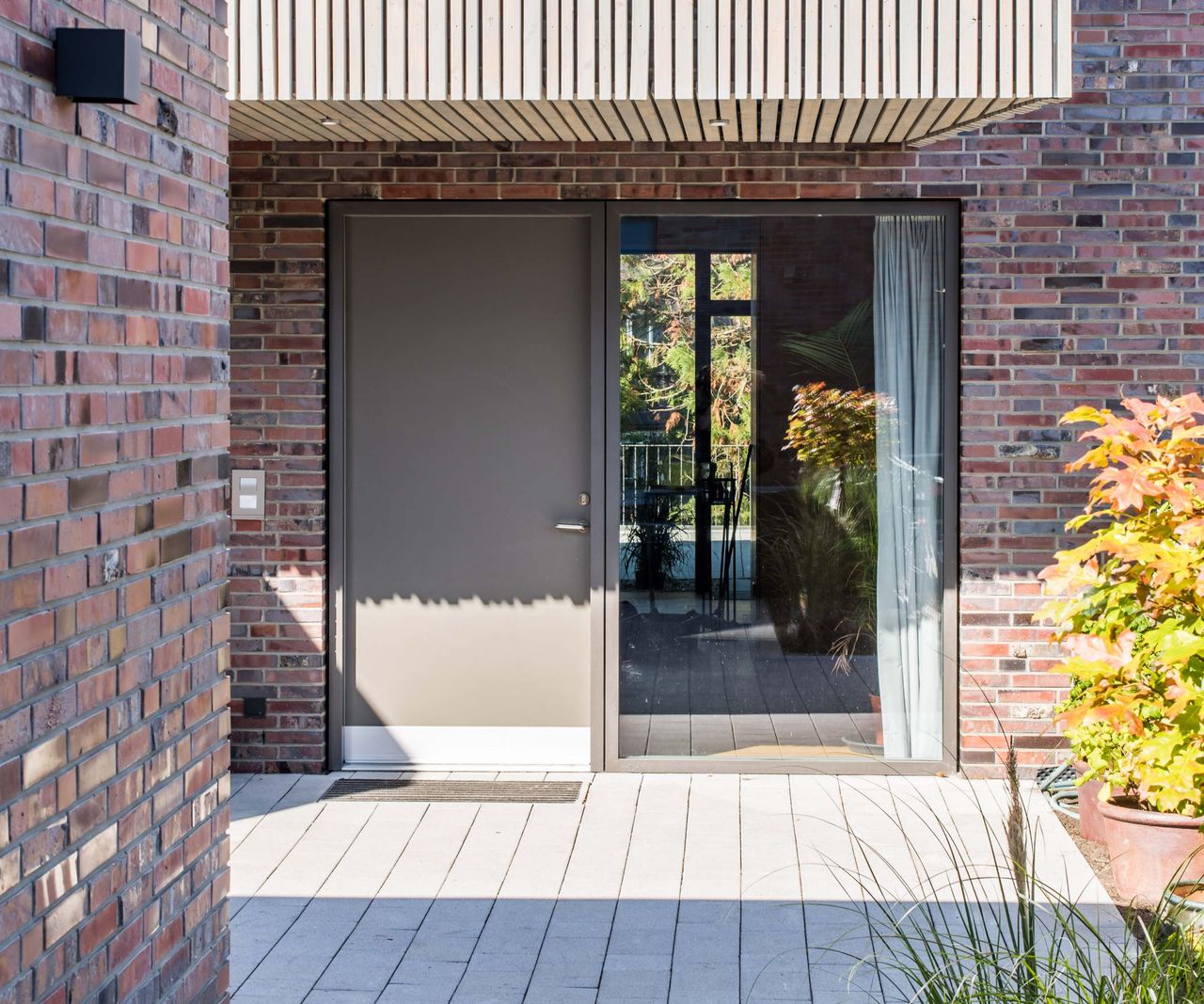 A close-up shot of a contemporary brick and timber-clad self build house showing a front door and adjacent fixed panel of glazing, from VELFAC, sat beneath a roof overhang.