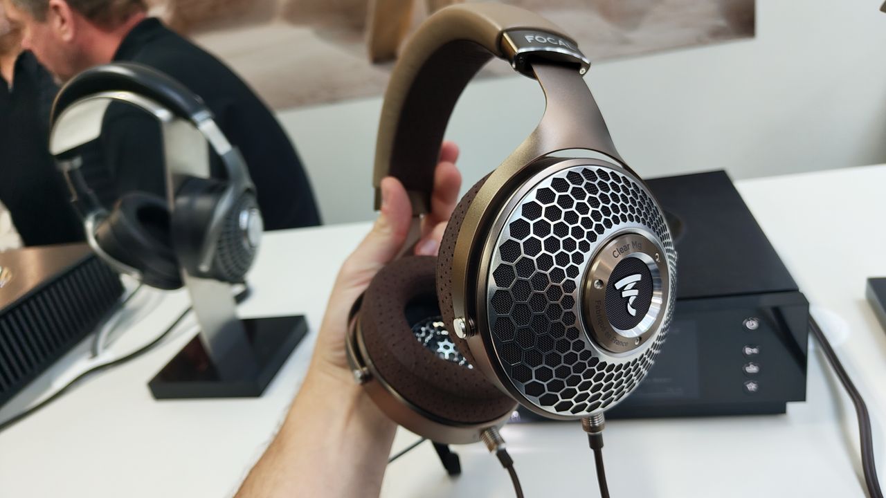 The Focal Clear Mg headphones held in the hand in front of a hi-fi component and another pair of headphones on a headphone stand.