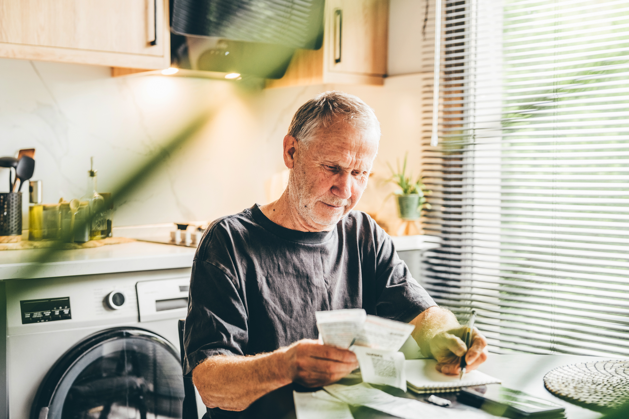 Man who has reached state pension age sits at kitchen table as he looks at paper documents and makes notes with a pen.