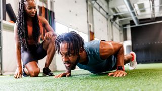 Man performs push-up in gym, a woman kneels beside him