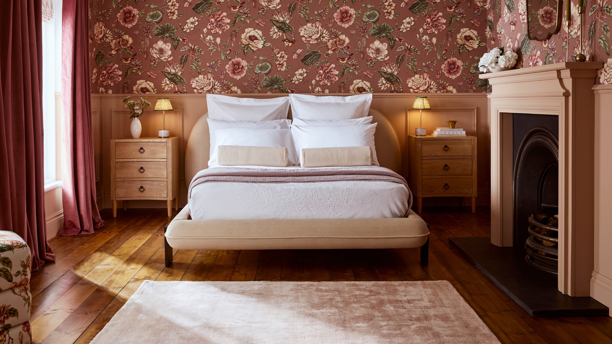 A pink bedroom with dado rail wall panelling and fireplace painted in Graham &amp; Brown's Rose Earth pink shade and the upper wall covered in the Roselle Grove wallpaper