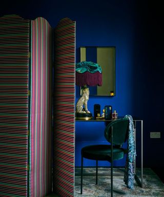 Corner of a bedroom with deep midnight blue walls, a dressing table with mirror, chair and room screen.