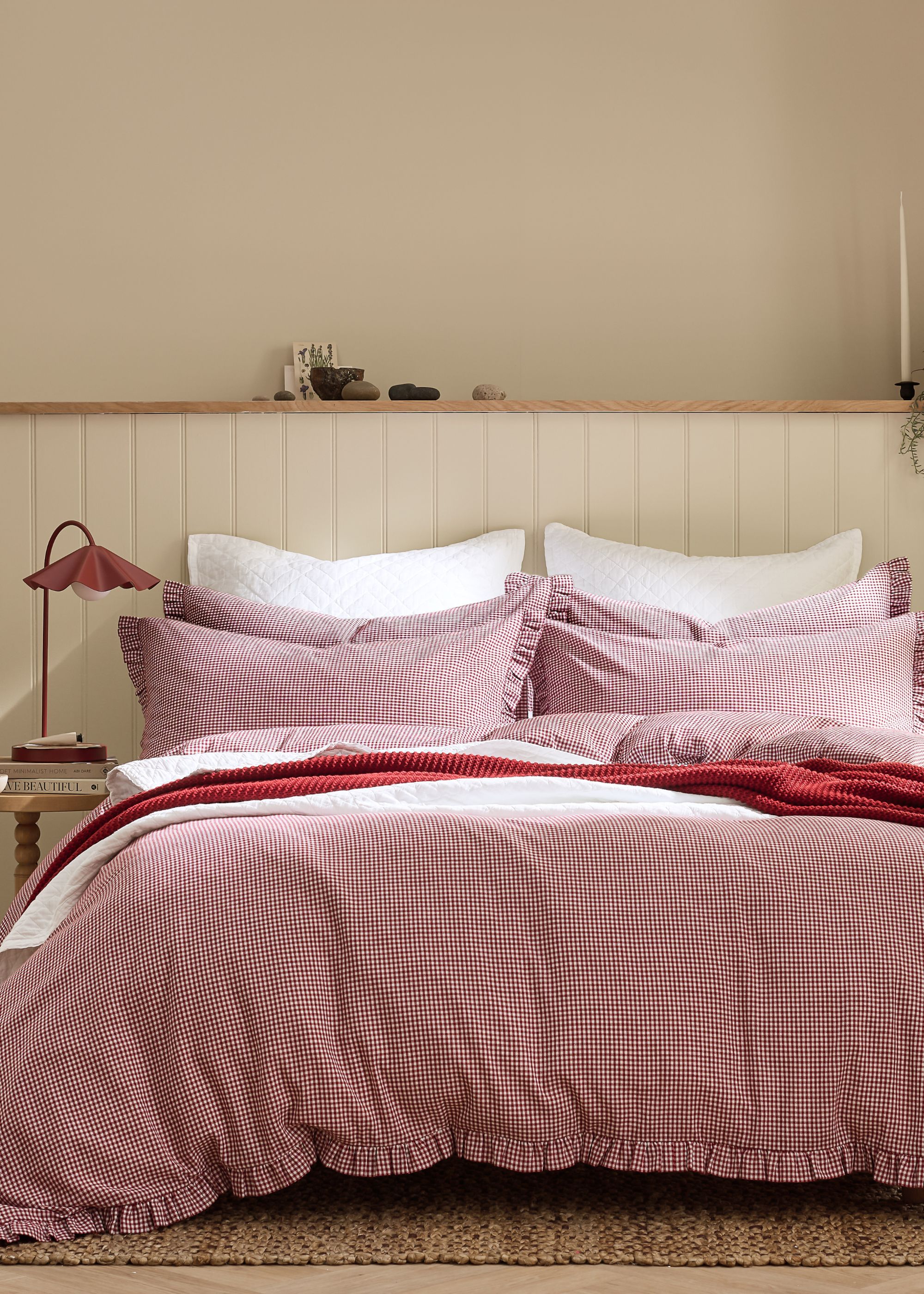 The corner of a bedroom with cream panelled walls, a double bed with red gingham bedding with white and red blankets layered on top.