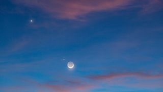 light blue sky with pink clouds and crescent moon with earthshine in the center with venus to the upper left and jupiter further to the upper left.