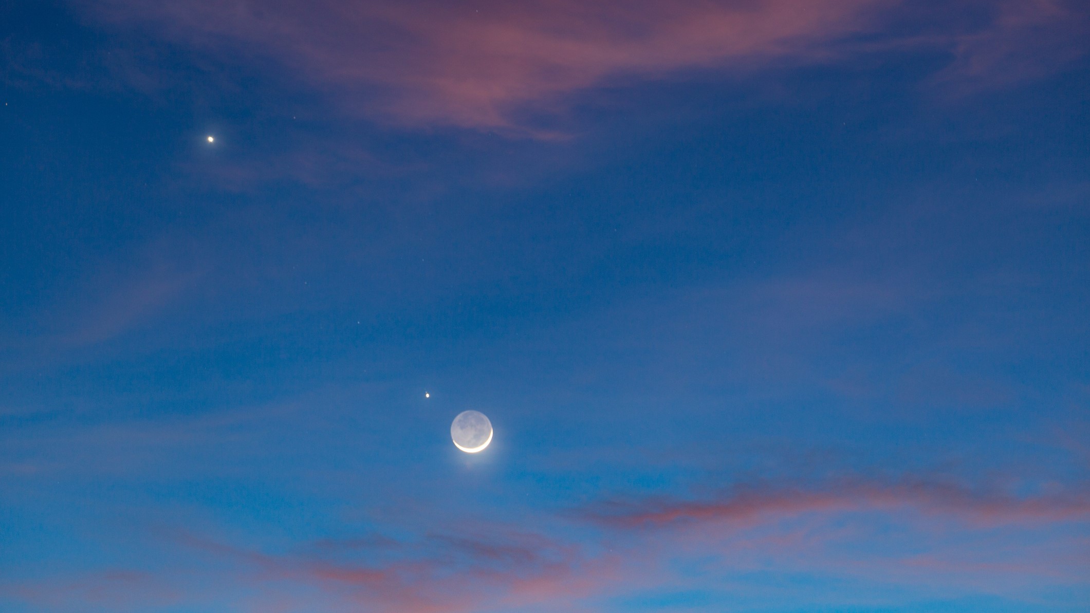 light blue sky with pink clouds and crescent moon with earthshine in the center with venus to the upper left and jupiter further to the upper left.