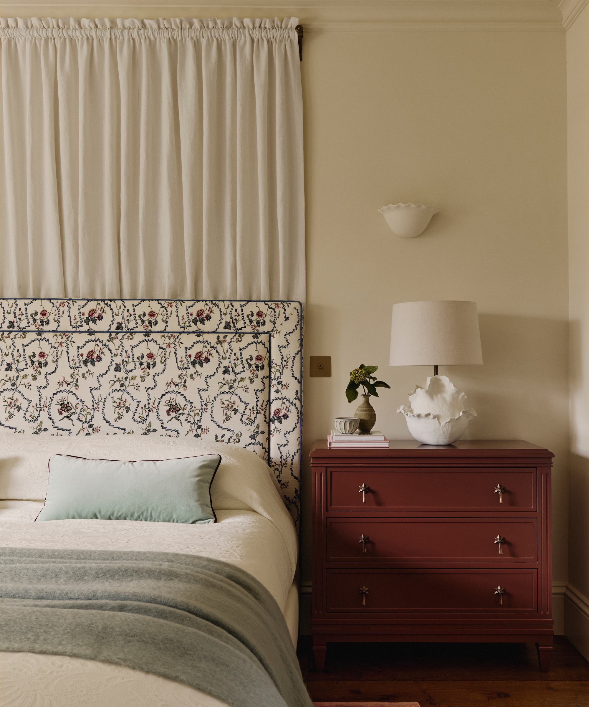 a bedroom with a patterned fabric headboard, a wooden bedside table, and a fabric curtain behind the bed