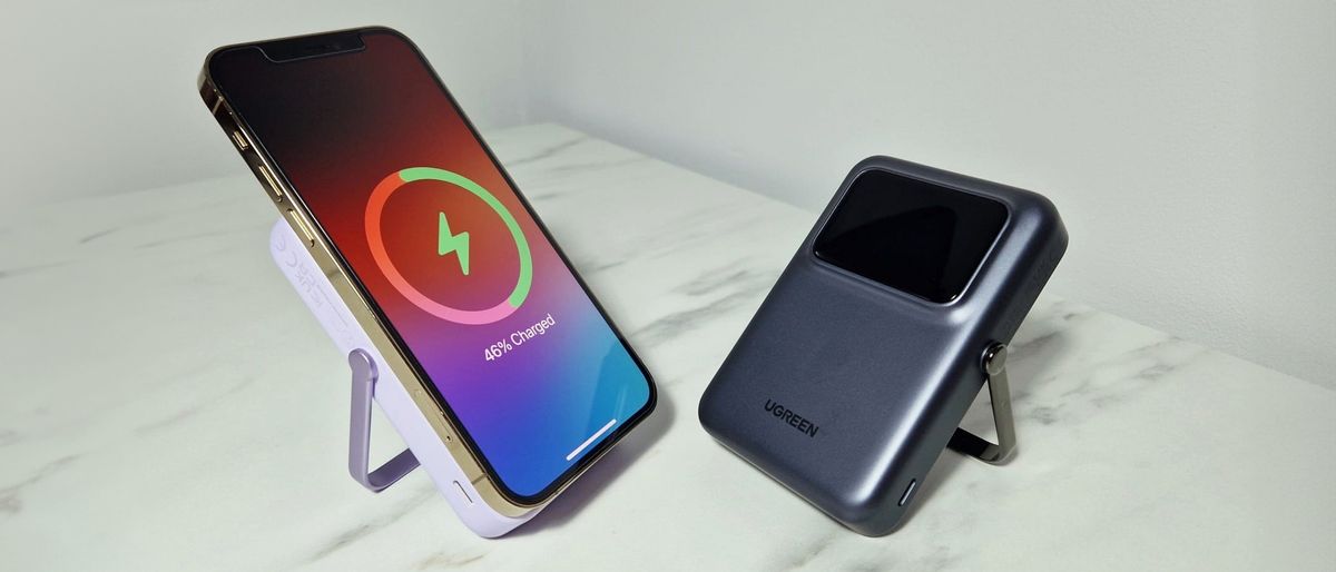 Two UGREEN Uno 15W Qi2 power banks are side by side on a marble effect surface, angled slightly toward each other. The power banks are gray and purple, the latter has an iPhone 12 Pro charging on it.