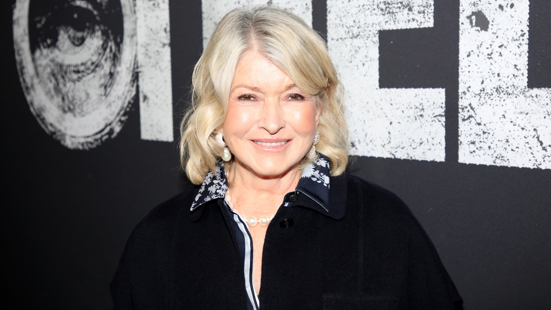 Image of Martha Stewart smiling with a curled blonde bob, a glowing complexion and wearing a navy shirt with white embroidery and a black jacket