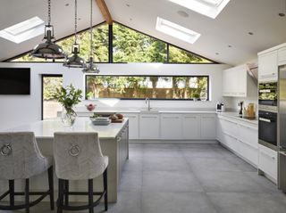 Harvey Jones Linear kitchen in a kitchen extension – an all-white kitchen with wide windows on the far wall, with a wide white kitchen island, velvet stools, and a grey slate floor, with hanging stainless steel lights