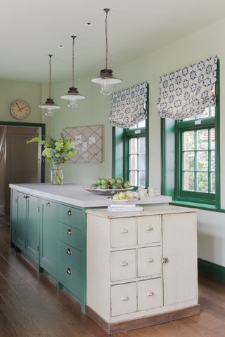 green kitchen with green island, green painted windows, vintage cabinet, trio of vintage pendant lamps, white countertop, blinds at windows