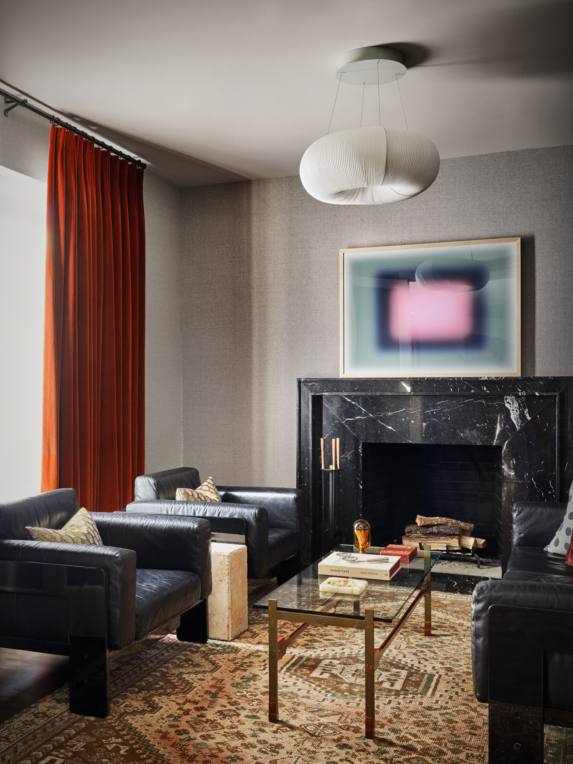 small living room with gray walls, a marble fireplace surround with artwork on top, red curtains, a patterned rug, two black leather armchairs, and a glass coffee table