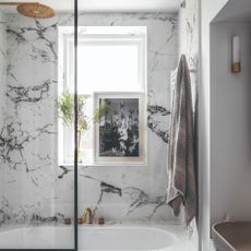 Bathroom with white and pale grey marbled walls and bath panel. Bath with gold coloured showerhead and window with a framed black and white photograph on the windowsill
