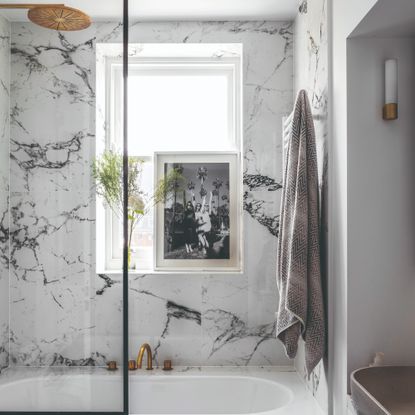 Bathroom with white and pale grey marbled walls and bath panel. Bath with gold coloured showerhead and window with a framed black and white photograph on the windowsill