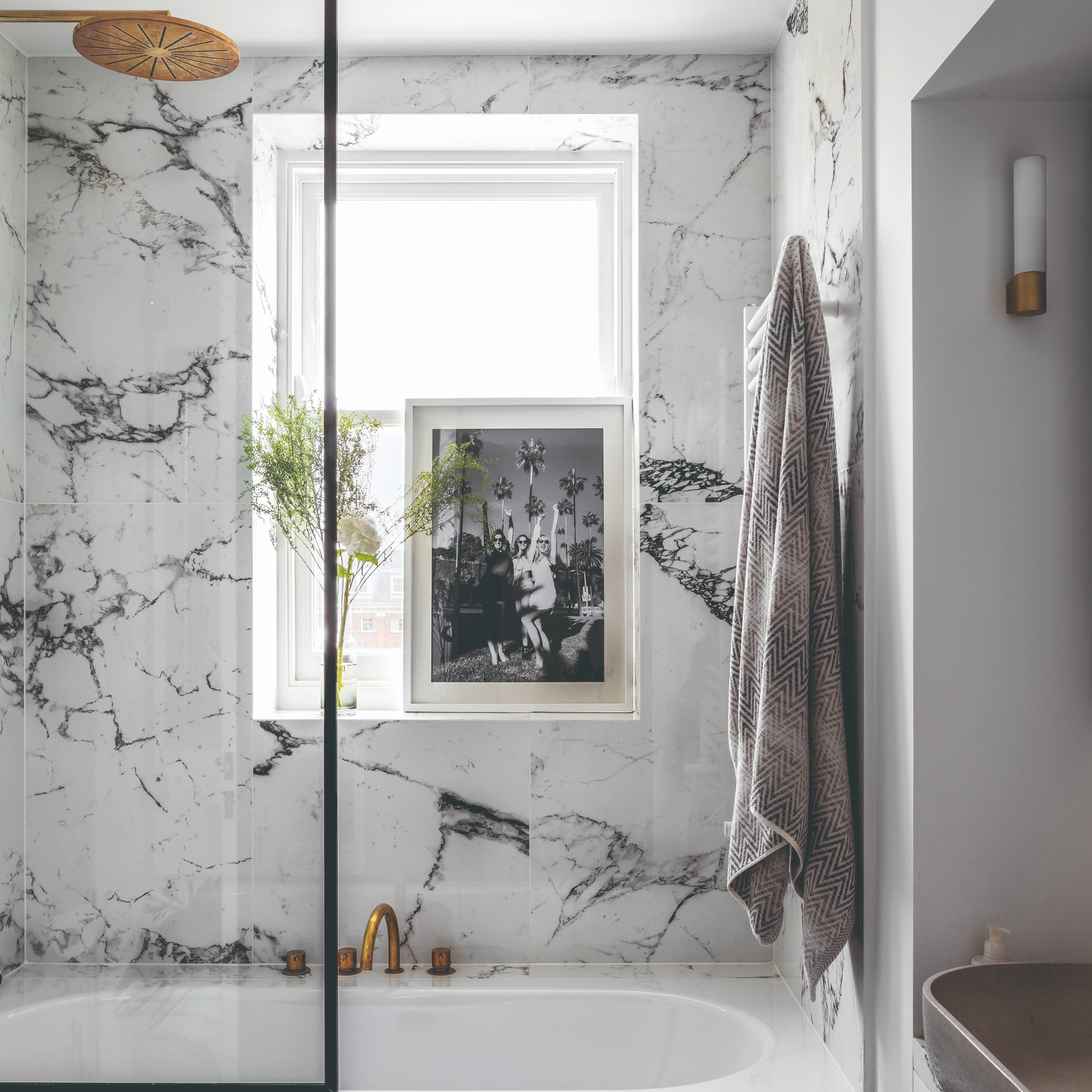 Bathroom with white and pale grey marbled walls and bath panel. Bath with gold coloured showerhead and window with a framed black and white photograph on the windowsill
