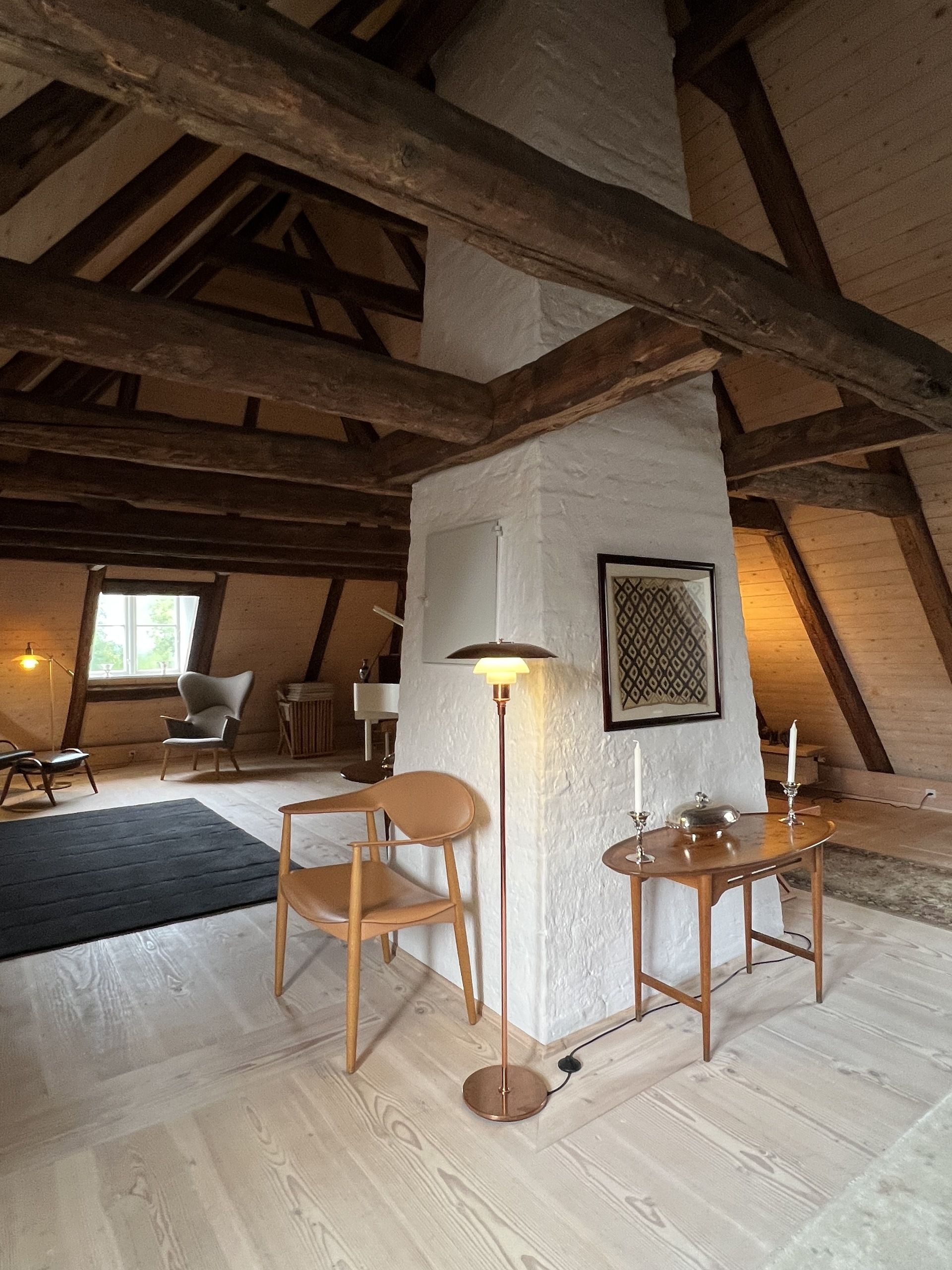 Image of a cottage style attic with old wood beams covering the A-frame ceiling. There are different pieces of Danish furniture around the room.