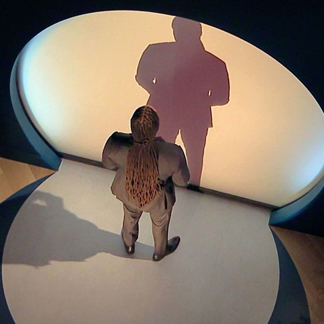 A man with long braided hair stands in front of a circle-shaped illuminated wall, in a sneak peek at 'Love is Blind' season 10.