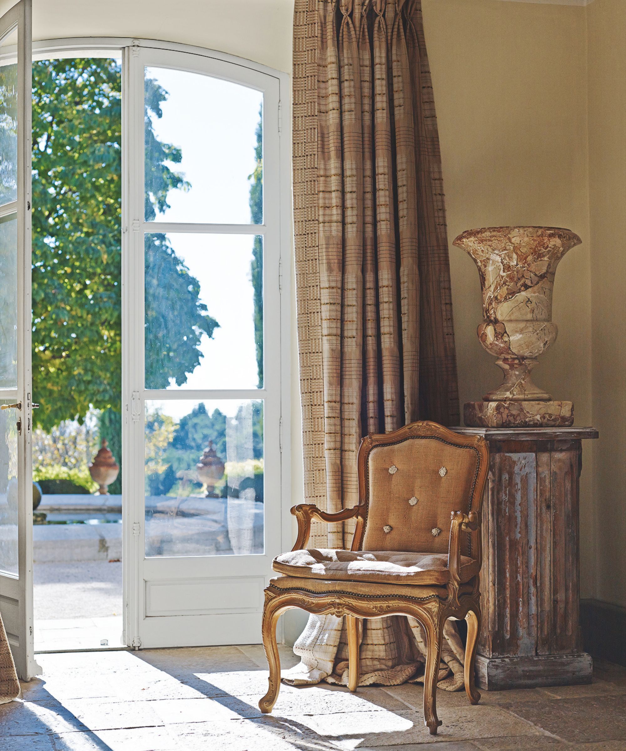 Hallway with ornate gold chair, pillar with marble vase, French doors onlooking garden