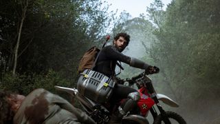 Manel riding a motorbike with a cat carrier on the back as a dead zombie lies on the floor in front of him in Apocalypse Z: The Beginning of the End.