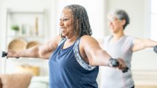 older black woman holding two dumbbells out to her sides at shoulder-height with another woman out of focus in the background. 