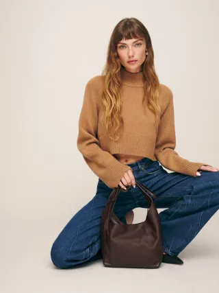 Model crouched wearing camel turtleneck, blue jeans, and brown handbag