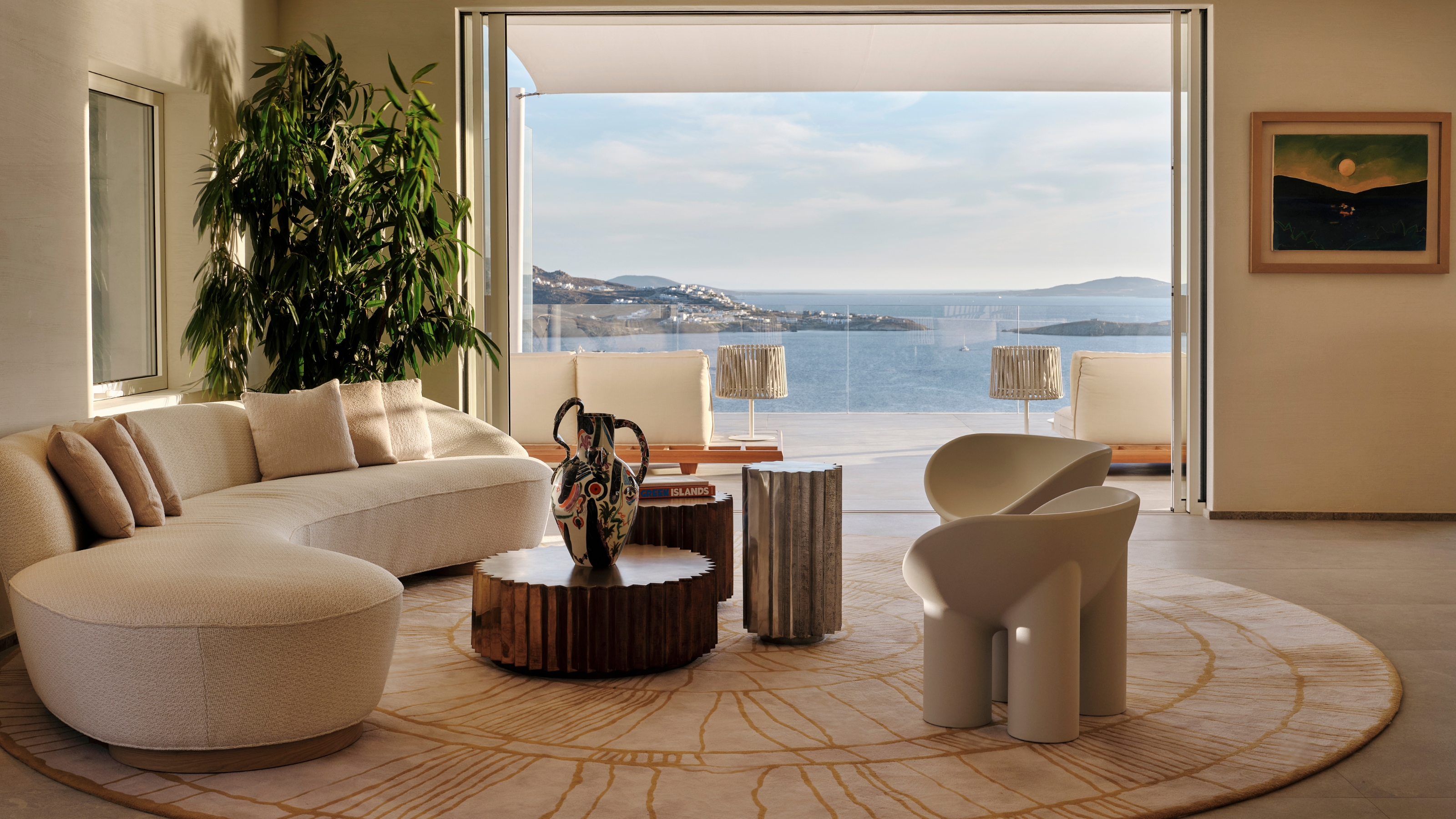 A sea-view hotel suite with sculptural contemporary furnishings including legged, one-piece plastic chairs that embrace the body&#039;s silhouettes, a sprawling beige sofa, ceramic vases, and wood and chrome coffee tables.